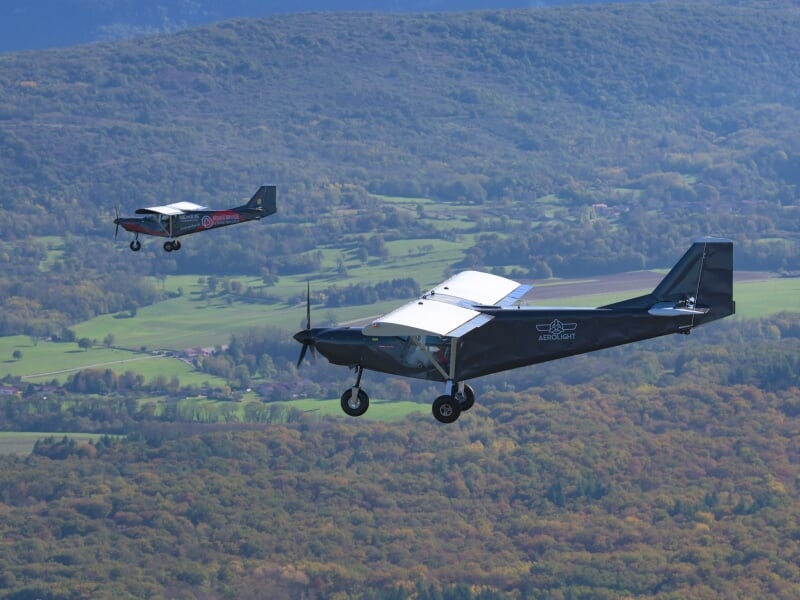Deux avions légers volant en formation au-dessus d'une vallée verdoyante, avec des montagnes boisées en arrière-plan.