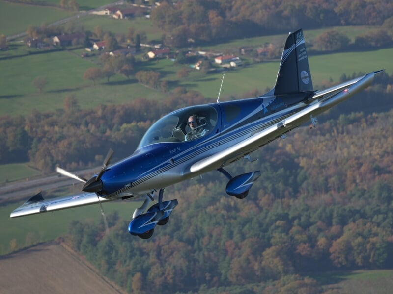 Avion léger bleu survolant une campagne verdoyante en arrière-plan, avec le pilote visible dans le cockpit. L'appareil est un ULM, modèle XL-8R, identifiable par ses lignes aérodynamiques et sa peintu