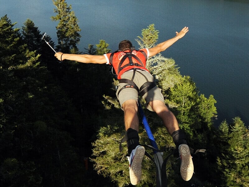 Homme en short et t-shirt rouge effectuant un saut à l'élastique au-dessus d'une forêt dense et d'un lac.