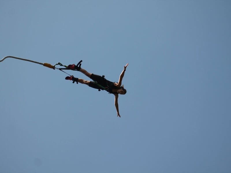 Femme en tenue noire effectuant un saut à l'élastique, bras écartés, suspendue dans un ciel bleu clair.