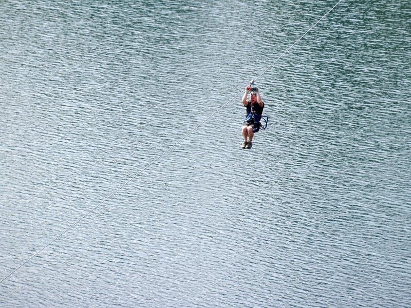 Personne suspendue à une tyrolienne au-dessus d'un lac calme, entouré de forêts verdoyantes.