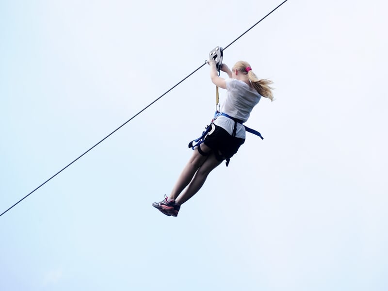 Femme blonde en short et t-shirt gris glissant sur une tyrolienne dans un ciel clair et lumineux.