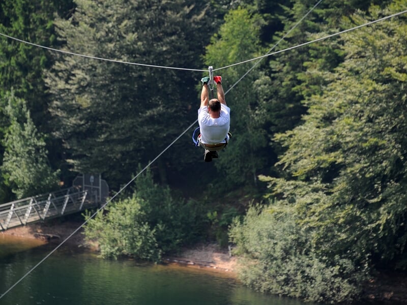 Homme en t-shirt blanc accroché à une tyrolienne au-dessus d'un lac, entouré d'arbres verts.