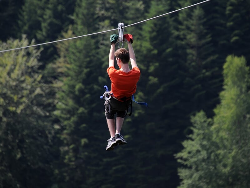 Jeune garçon en t-shirt orange suspendu à une tyrolienne au-dessus d'un lac et d'une forêt dense.
