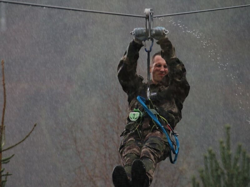 Personne en tenue camouflage souriante, suspendue à une tyrolienne dans un environnement naturel boisé.