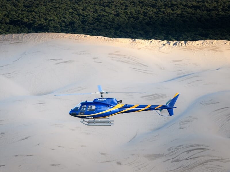 Hélicoptère évoluant au-dessus des grandes dunes de sable et d’une forêt dense, soulignant un paysage naturel spectaculaire et préservé