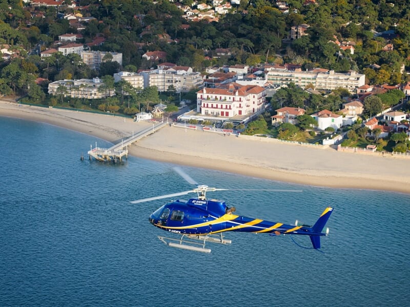 Hélicoptère bleu et jaune survolant une plage de sable fin bordée de villas et d'un ponton, entourée de végétation et d'habitations élégantes