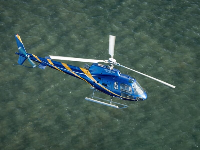 Vue plongeante d’un hélicoptère volant au-dessus d’une mer calme et verdâtre, ses couleurs vives contrastant avec l’eau en contrebas