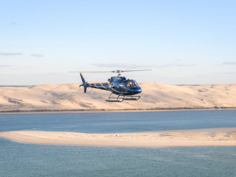 Hélicoptère volant près de bancs de sable et de la Dune du Pilat, survolant une eau turquoise dans un paysage naturel spectaculaire