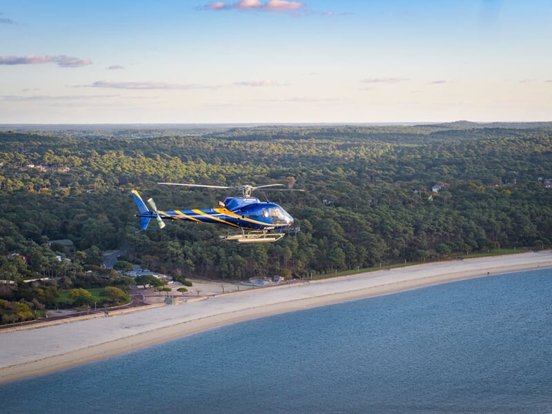 Hélicoptère bleu et jaune survolant une plage sablonneuse avec forêt verdoyante en arrière-plan sous un ciel dégagé