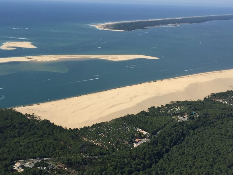 Vue aérienne de la Dune du Pilat bordée par une forêt dense et des bancs de sable entourés d’eau bleue calme