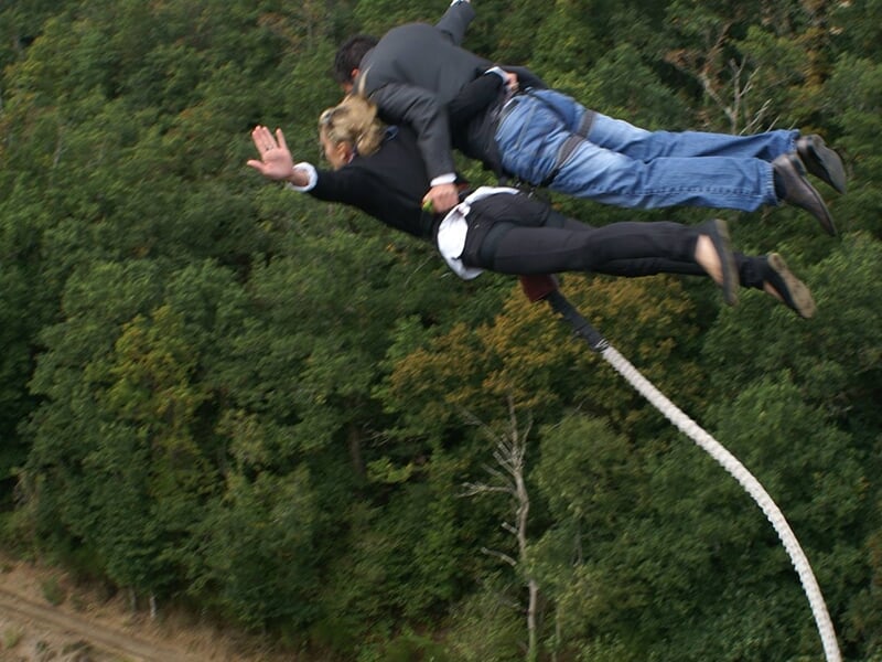 Couple en plein saut à l'élastique, suspendu dans les airs au-dessus d'une forêt dense et verdoyante.