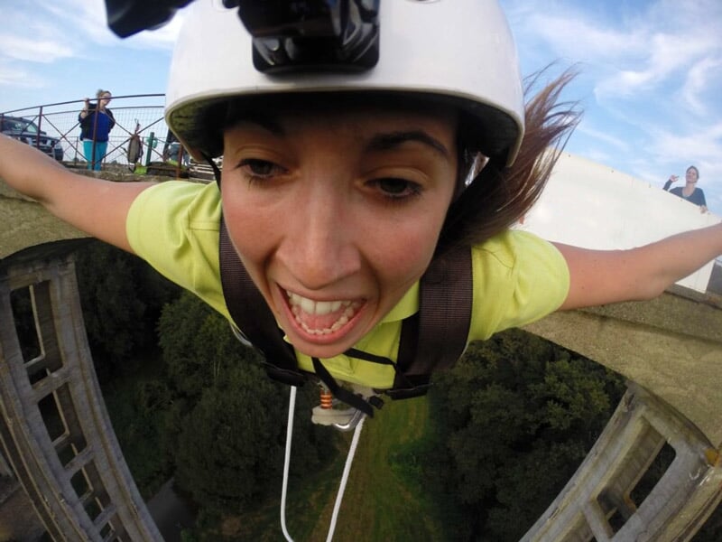 Gros plan sur le visage d'une femme criant de joie ou d'adrénaline durant un saut à l'élastique, portant un casque et un harnais, avec une vue en arrière-plan d'un pont et d'une végétation dense