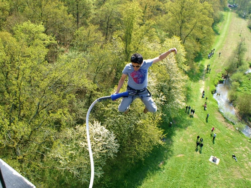 Homme en pleine descente lors d'un saut à l'élastique, équipé d'un harnais, au-dessus d'une campagne arborée avec des spectateurs visibles au sol