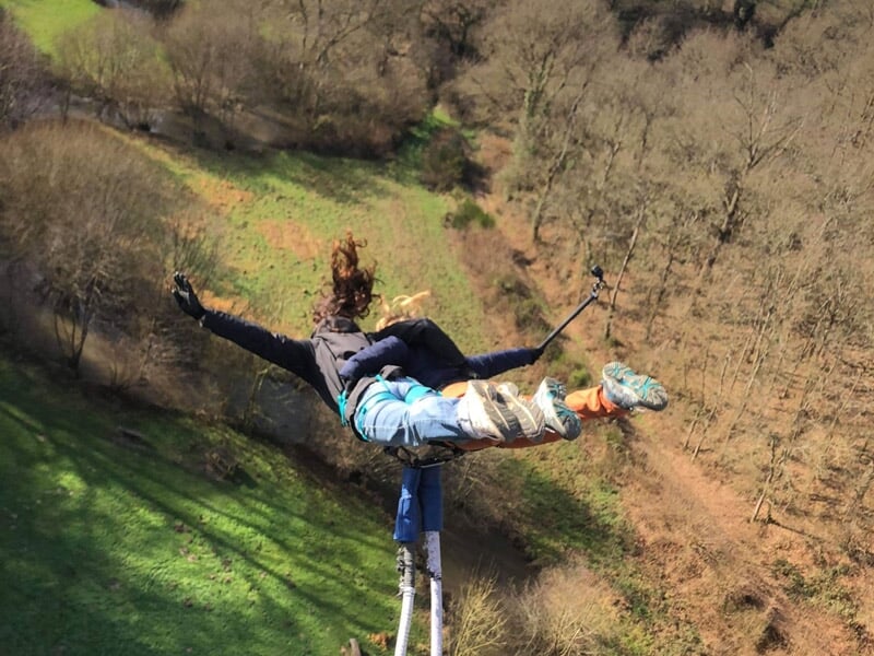 Vue plongeante sur une personne en plein saut à l'élastique, cheveux au vent, survolant une vallée aux tons verts et bruns, tenant une perche pour capturer le moment