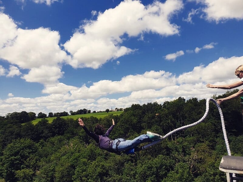 Saut à l'élastique d'une personne au-dessus d'une forêt dense, bras écartés, avec un ciel bleu parsemé de nuages blancs, et un instructeur visible sur la plateforme