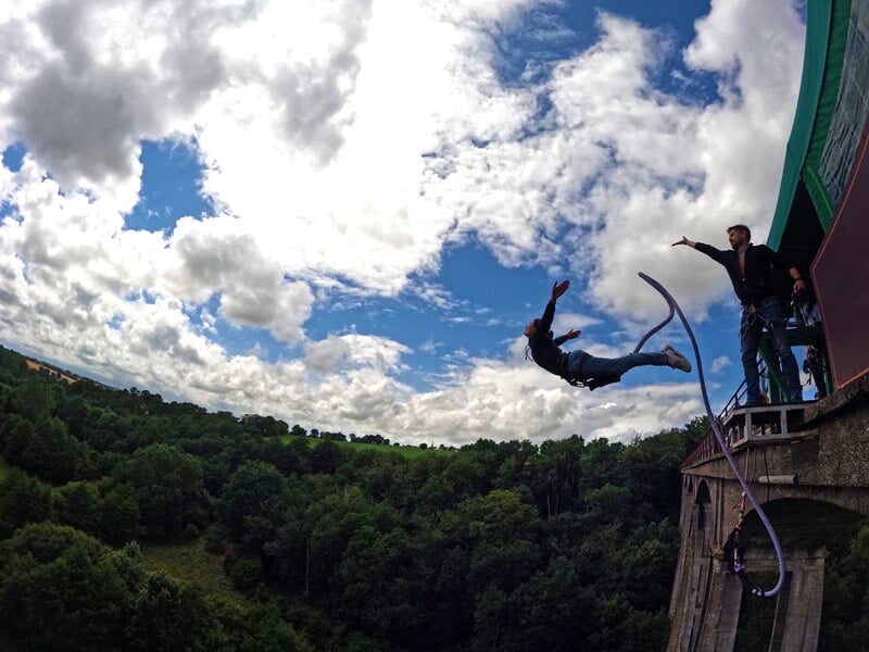 Personne effectuant un saut à l'élastique depuis un pont, entourée d'une vallée verdoyante et d'un ciel partiellement nuageux, avec un instructeur tenant la corde de sécurité