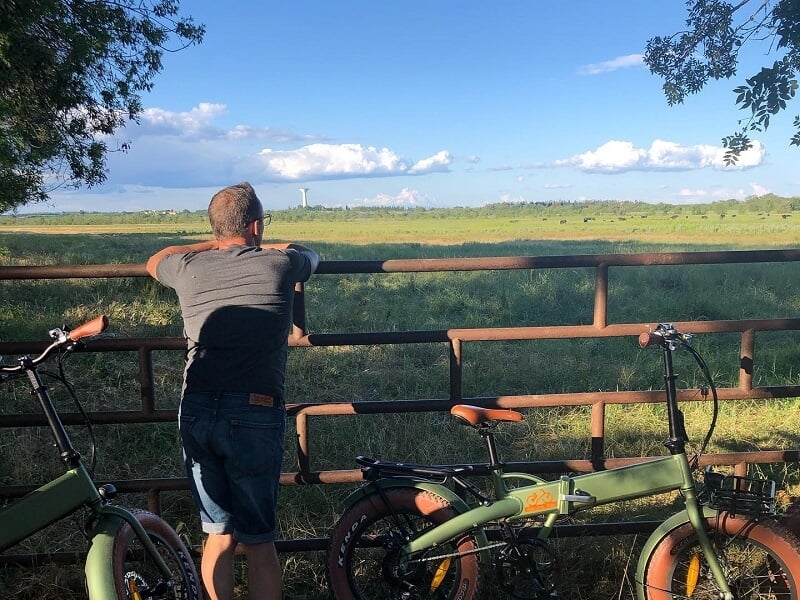 Homme appuyé sur une barrière en métal regardant un paysage vert avec deux vélos électriques garés à côté.