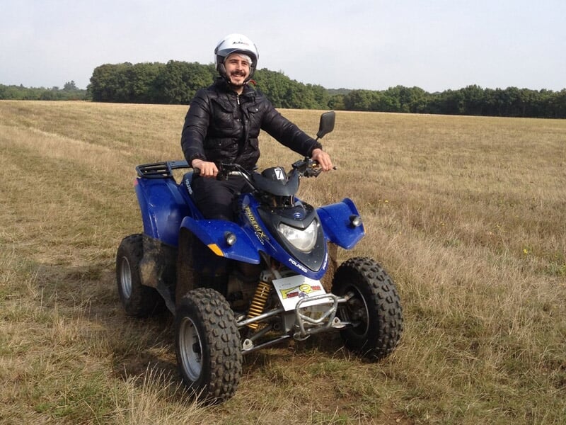 Homme souriant en casque assis sur un quad bleu dans un champ ouvert avec des arbres au loin.