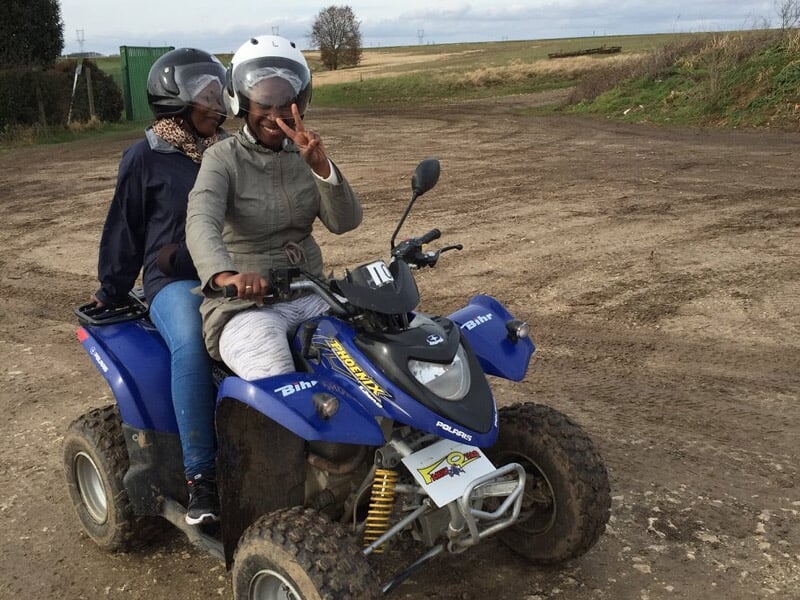 Deux personnes en casque sur un quad bleu sur un terrain de terre battue avec un ciel nuageux.