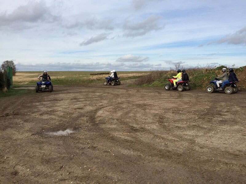 Quatre quads avec conducteurs en casque roulant sur un terrain plat et dégagé sous un ciel nuageux.