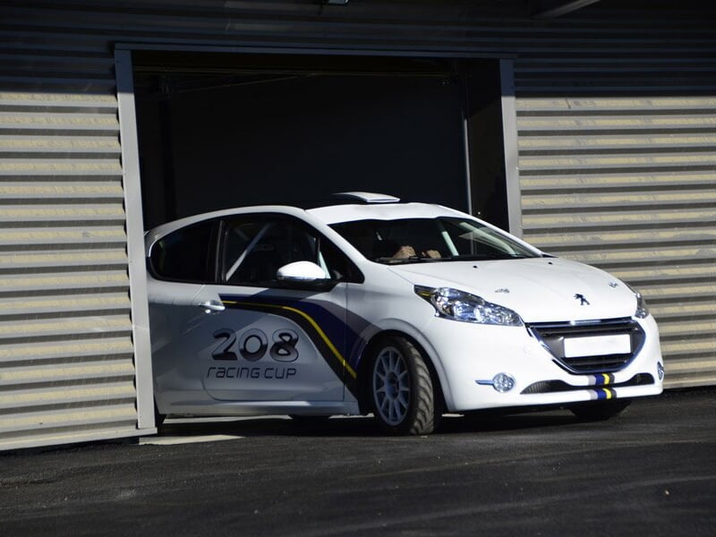 Voiture de course Peugeot 208 Racing Cup blanche sortant d'un garage sur un circuit d'asphalte en plein jour.