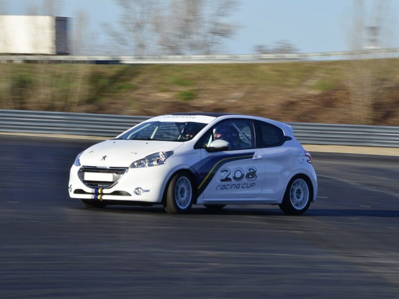 Voiture rouge de course sur circuit asphalte, virage serré avec deux pilotes casqués à bord.