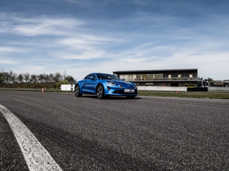 Alpine A110 bleue sur circuit d'asphalte à Andrézieux, sous un ciel partiellement nuageux, avec bâtiment en arrière-plan.