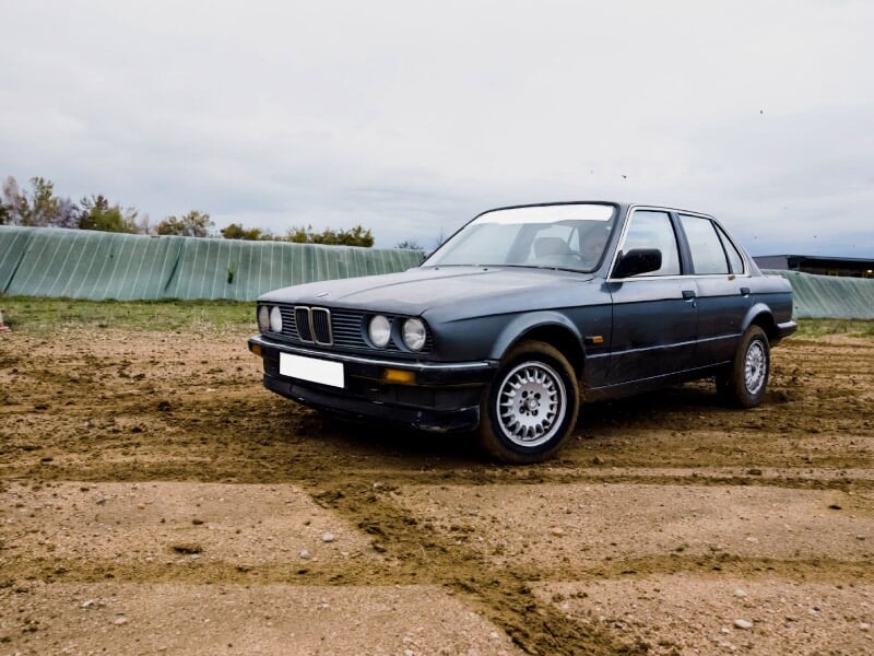 Voiture de rallye blanche sur circuit terre, virage serré avec poussière soulevée.