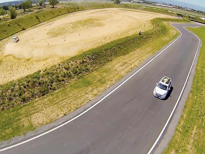 Voiture de rallye blanche sur circuit terre, virage serré avec poussière visible.