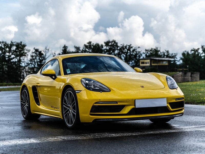 Voiture de sport jaune Porsche 718 Cayman S garée sur circuit humide avec ciel nuageux et arbres en arrière-plan.