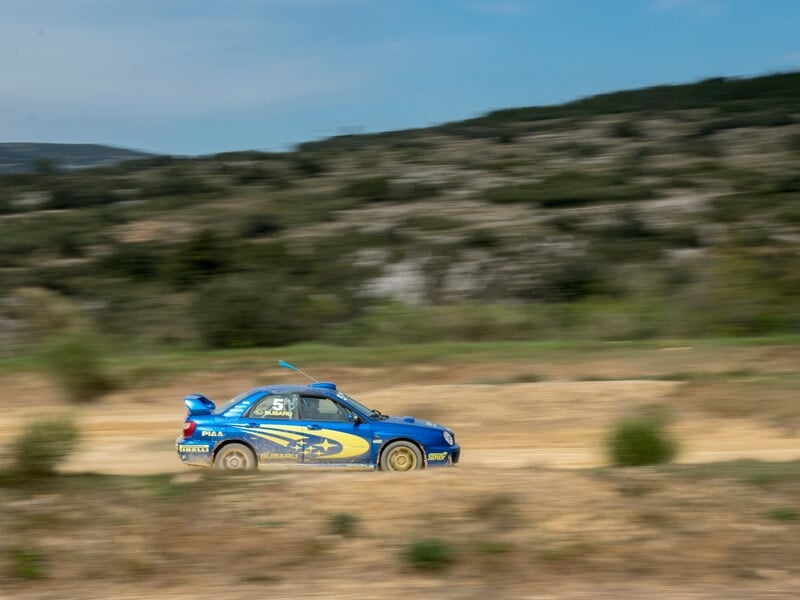 Voiture de rallye bleue en pleine course sur un circuit terre avec paysage naturel en arrière-plan flou.