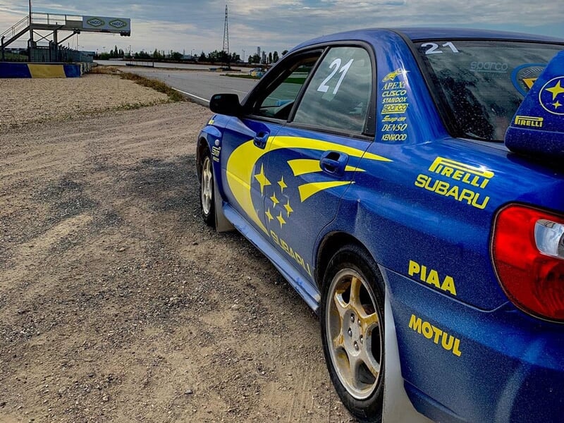 Vue rapprochée d'une voiture de rallye bleue stationnée sur un sol en gravier avec des logos de sponsors visibles.