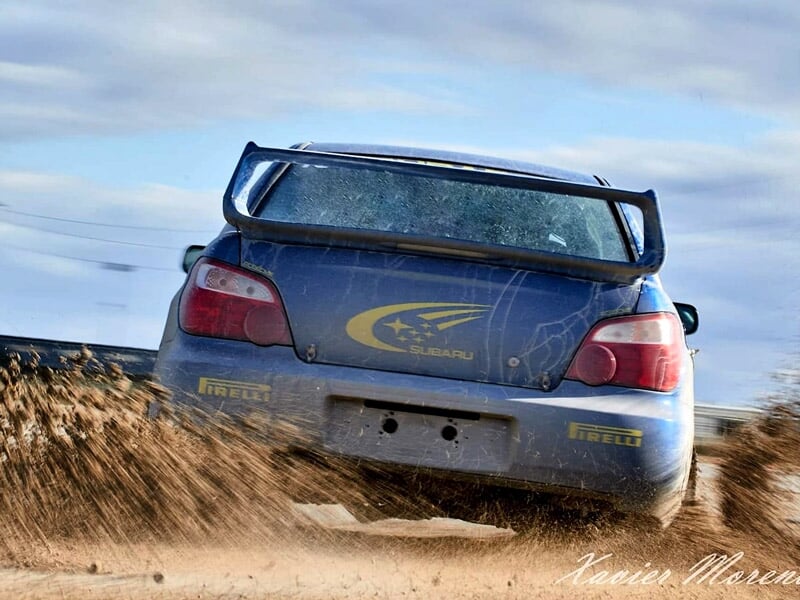 Arrière d'une voiture de rallye bleue soulevant de la poussière sur un circuit terre, ambiance dynamique et sportive.
