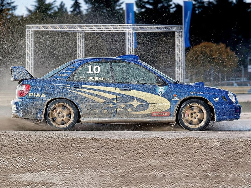 Voiture de rallye bleue en mouvement sur un circuit terre, éclaboussant de la boue avec un cadre métallique en arrière-plan.