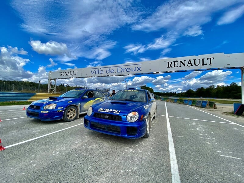 Deux voitures de rallye bleues alignées sur une piste bitumée sous un ciel bleu avec un panneau Renault Ville de Dreux.
