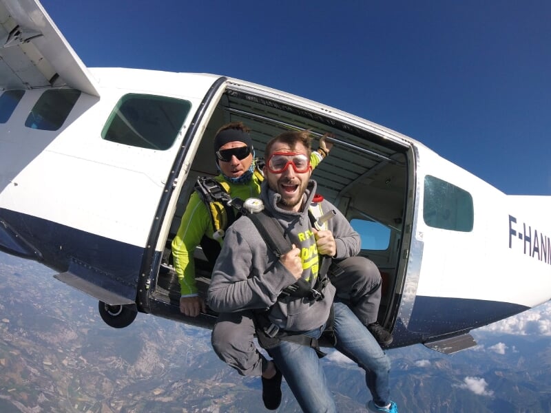 Homme souriant prêt à sauter en parachute depuis un avion blanc, accompagné d'un instructeur en arrière-plan.