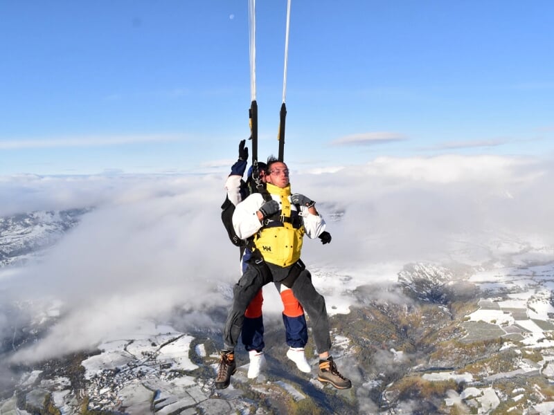 Deux parachutistes en tandem flottant au-dessus d'un paysage enneigé et nuageux, en pleine descente.