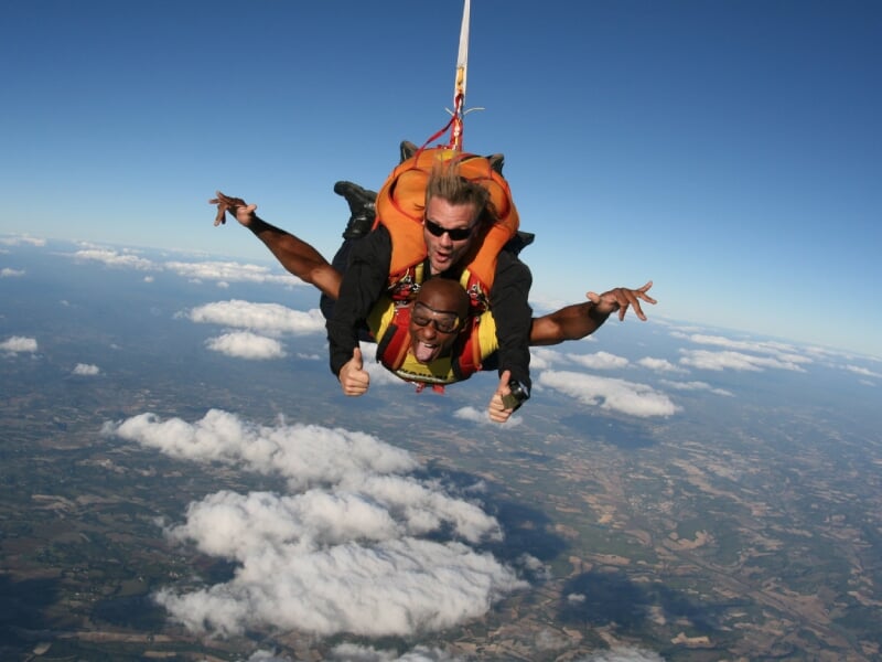 Saut en parachute en tandem avec un instructeur, les deux bras écartés, survolant un paysage avec des nuages.