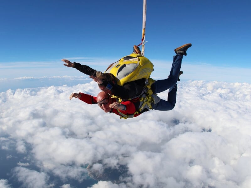 Parachutistes en tandem en chute libre au-dessus d'un ciel bleu et nuageux, avec un instructeur portant un sac jaune.