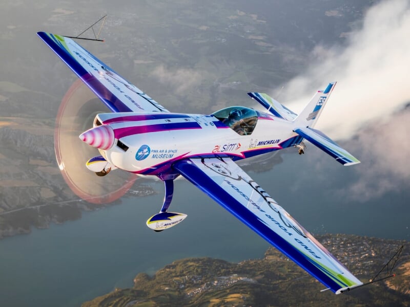 Avion de voltige coloré en vol au-dessus d'un lac et d'un paysage montagneux, avec une hélice en mouvement.