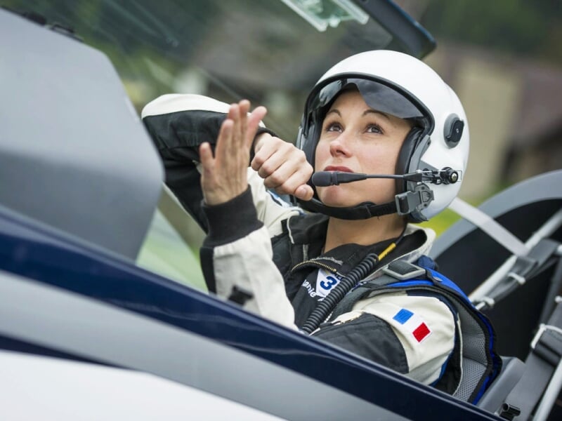Pilote féminine en combinaison et casque blanc, concentrée dans le cockpit d'un avion de voltige.
