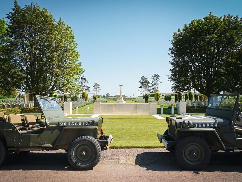 Deux jeeps militaires vintage face à face devant un cimetière militaire avec une grande croix au centre.