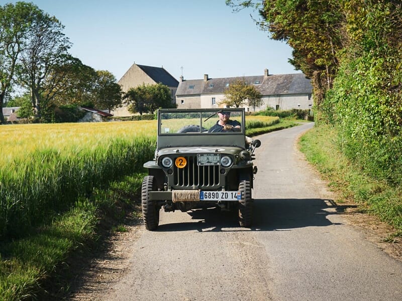 Une jeep militaire vintage conduite sur une route de campagne bordée de champs et de maisons en arrière-plan.