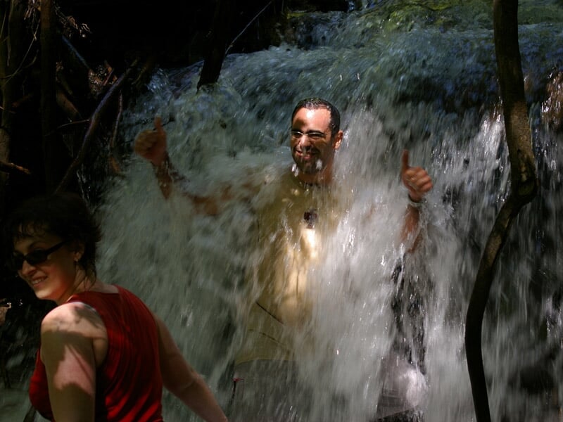 Homme souriant sous une cascade d'eau en forêt, accompagné d'une femme en débardeur rouge.