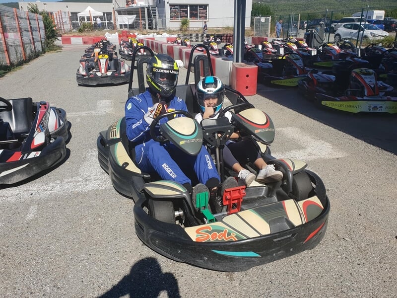 Deux personnes portant un casque sont assises dans un kart sur une piste de karting en extérieur par une journée ensoleillée.