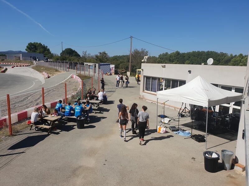 Des personnes marchent près d'une tente blanche et des tables sur un circuit de karting avec une piste visible à gauche.