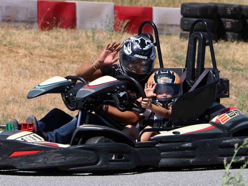 Un adulte et un enfant portant un casque dans un kart double sur une piste de karting, l'enfant fait un signe de la main.