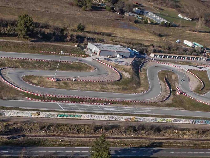 Vue aérienne d'un circuit de karting sinueux entouré de végétation et de quelques bâtiments en arrière-plan.