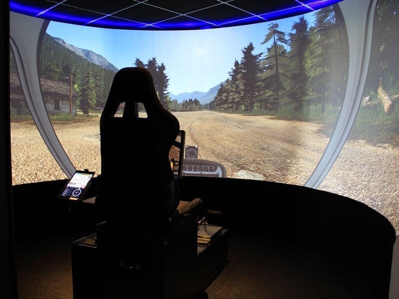 Simulateur de pilotage d'avion avec siège et grand écran circulaire montrant un décor naturel de forêt et montagne.